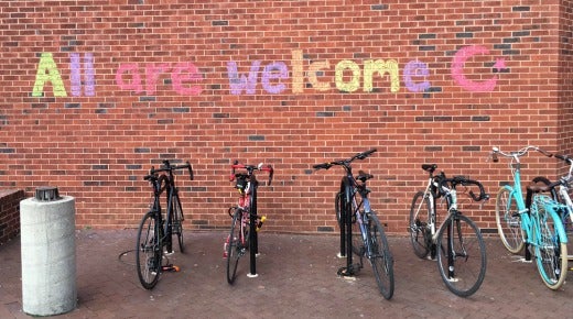 Bicycles parked outside brick building with the words written above them &quot;All are welcome&quot;